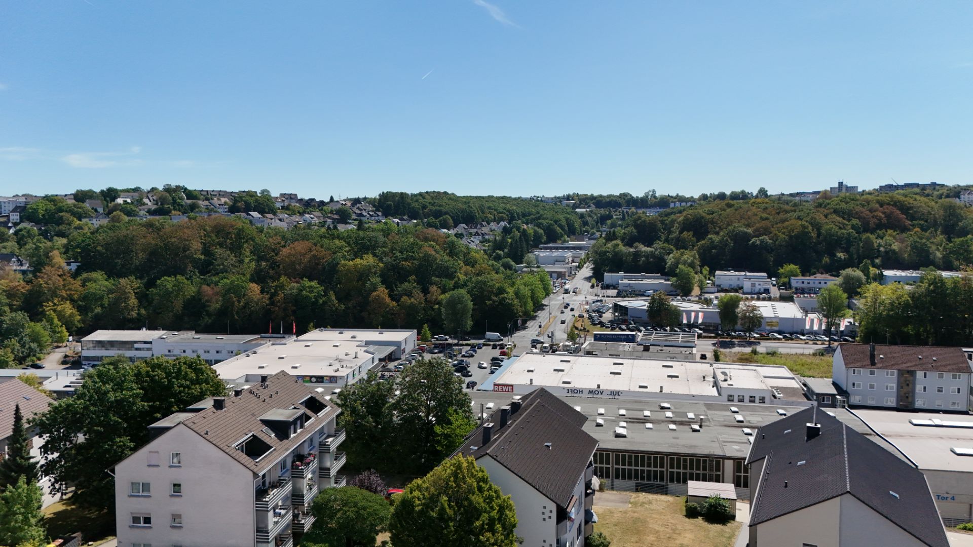 Blick auf Wohnhäuser, Gewerbegebäude und eine von Bäumen gesäumte Straße bei sonnigem Wetter.