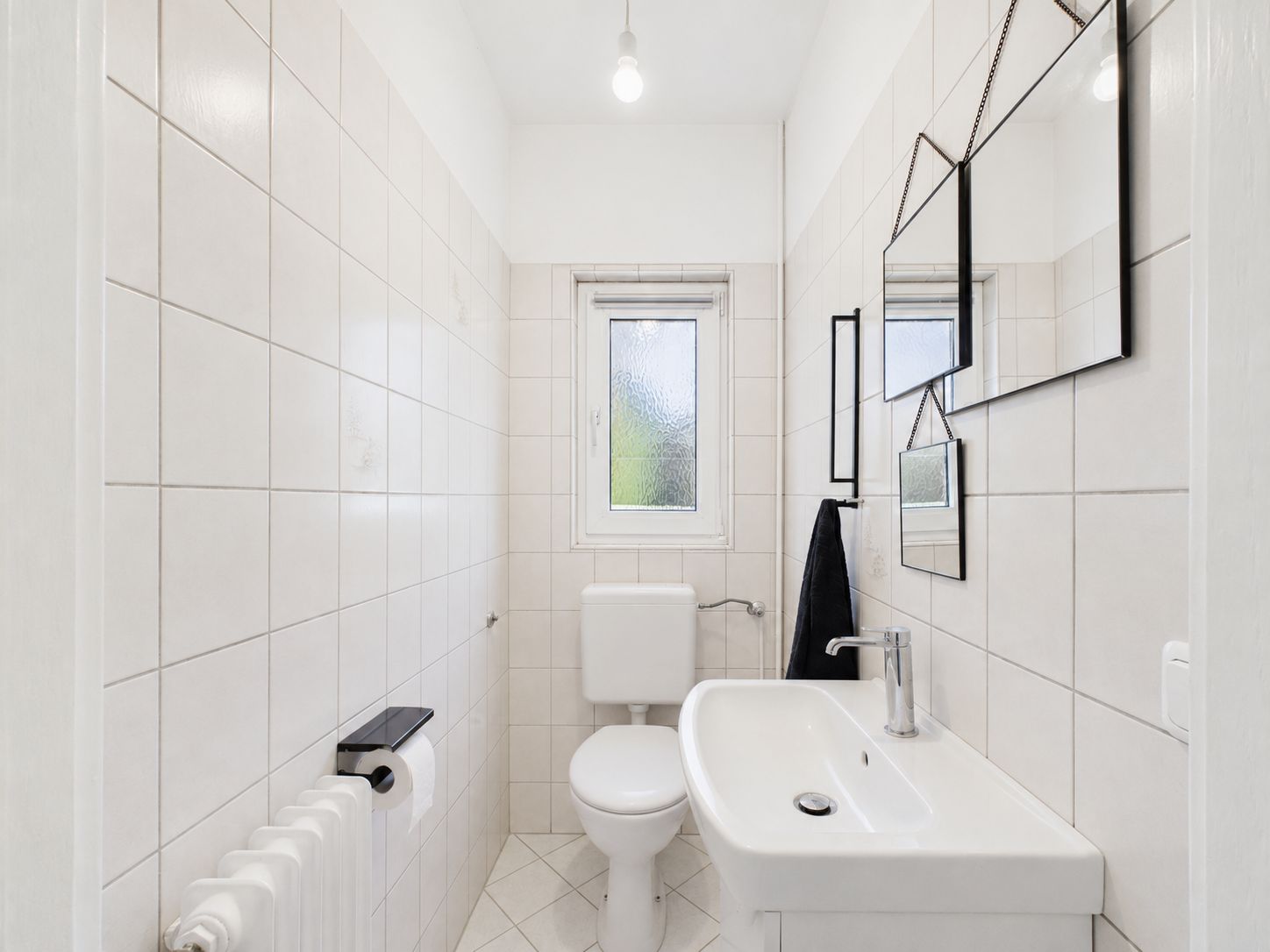 Helles, gefliestes Badezimmer mit Toilette, Waschbecken, Spiegel und Fenster.