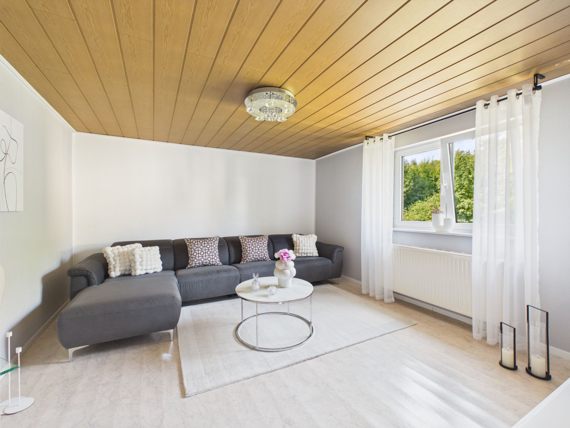 Helles Wohnzimmer mit grauem Ecksofa, rundem Tisch und großem Fenster mit weißen Vorhängen.