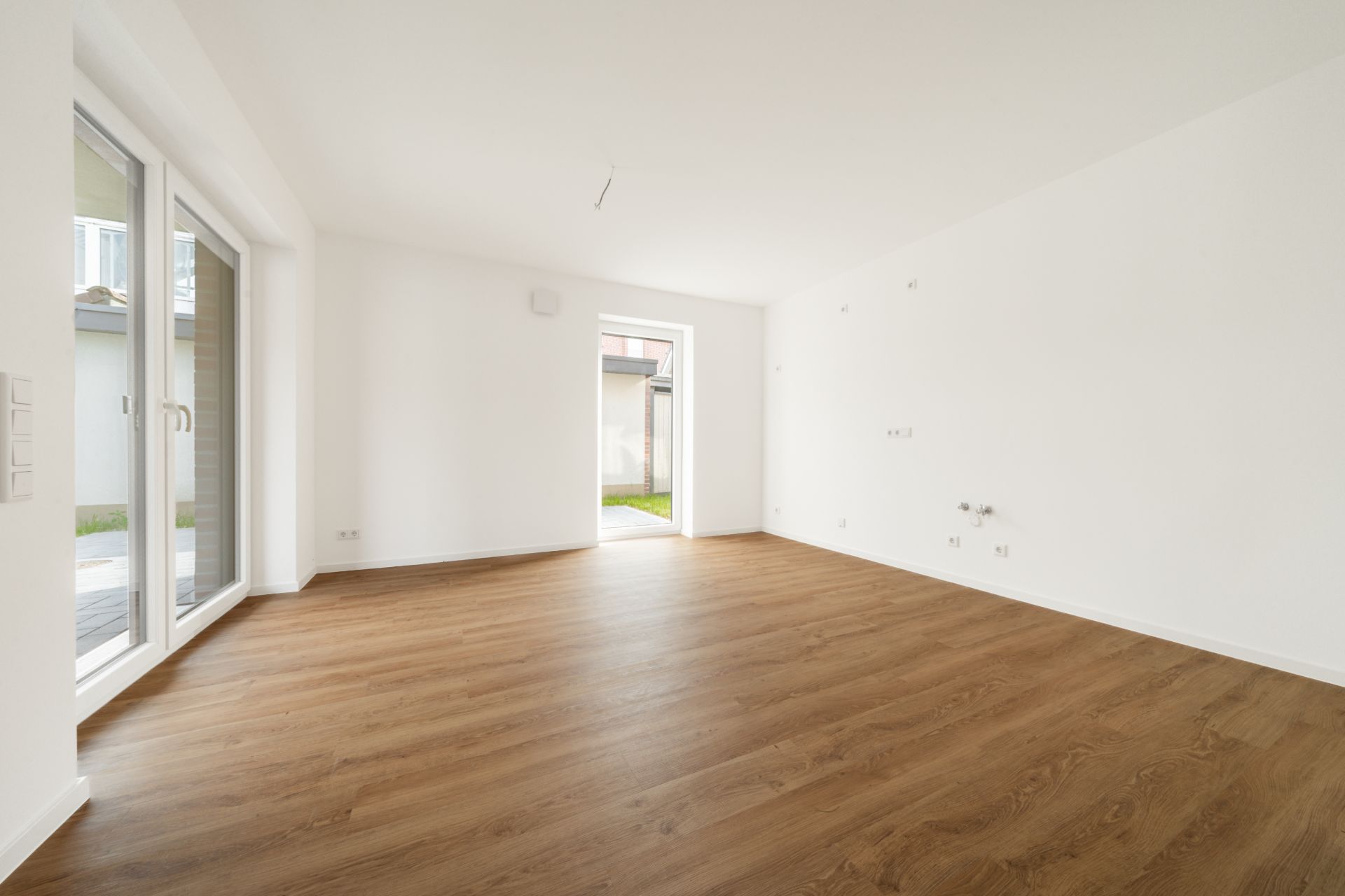 Leerer Raum mit Holzfußboden, weißen Wänden und großen Fenstern zur Terrasse.