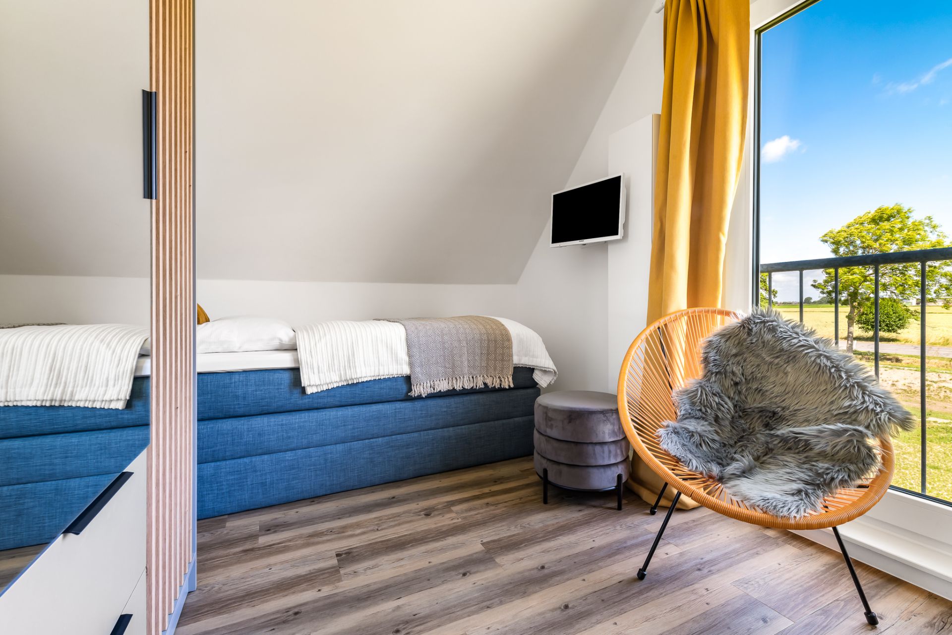 Modernes Schlafzimmer mit blauem Bett, Sessel, kleinem TV und großem Fenster mit Ausblick.