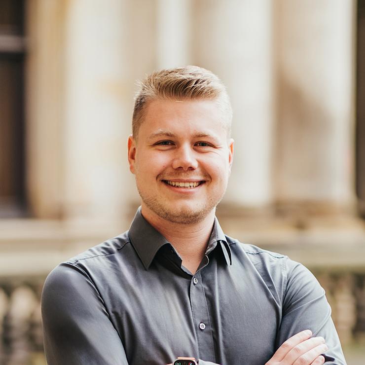 Ein lächelnder Mann steht mit verschränkten Armen vor einem Gebäude.