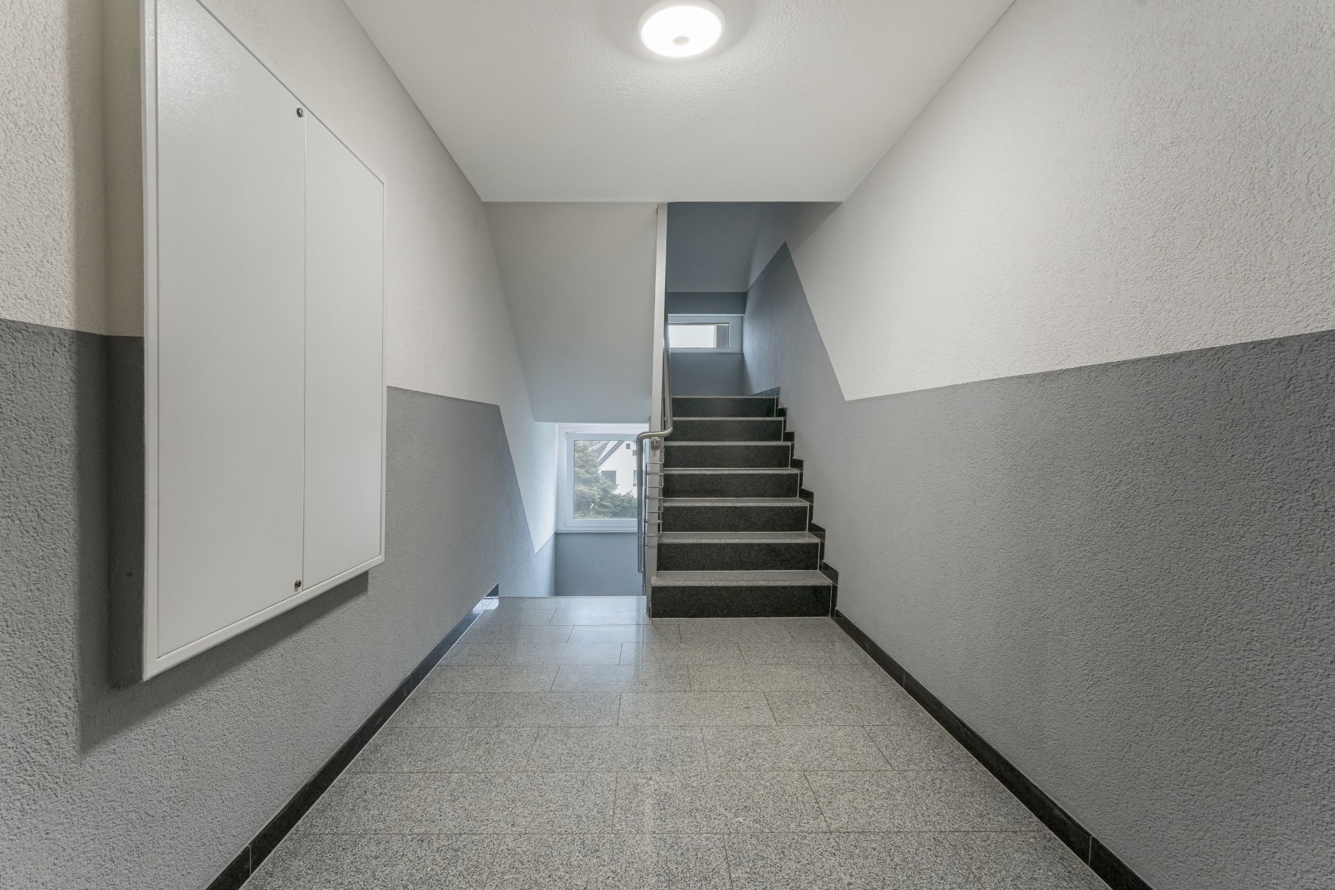 Helles Treppenhaus mit grauen Fliesen und Fenster am Ende der Treppe.