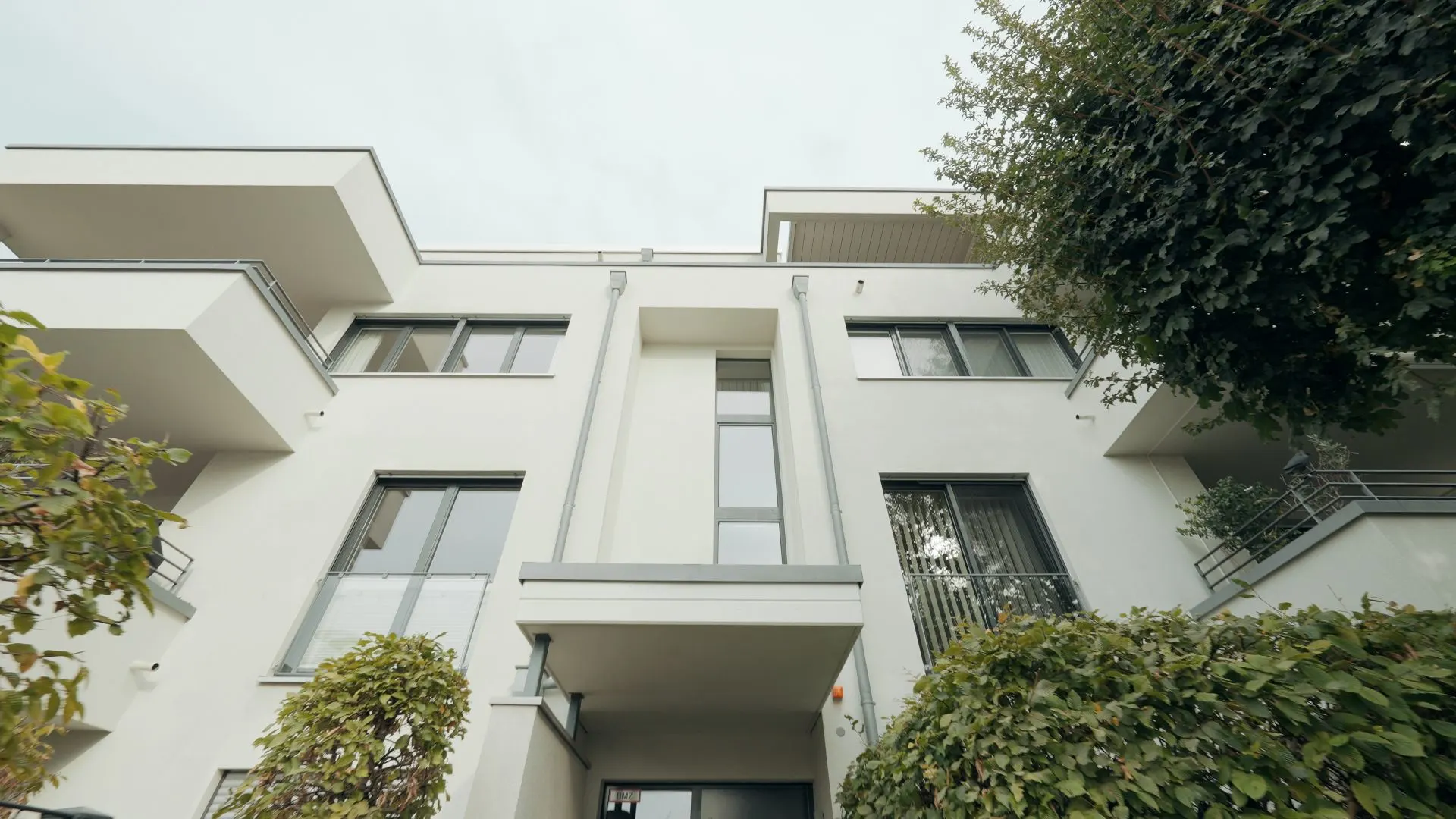 Moderne, weiße Wohnhausfassade mit großen Fenstern, Balkonen und umliegenden Büschen.
