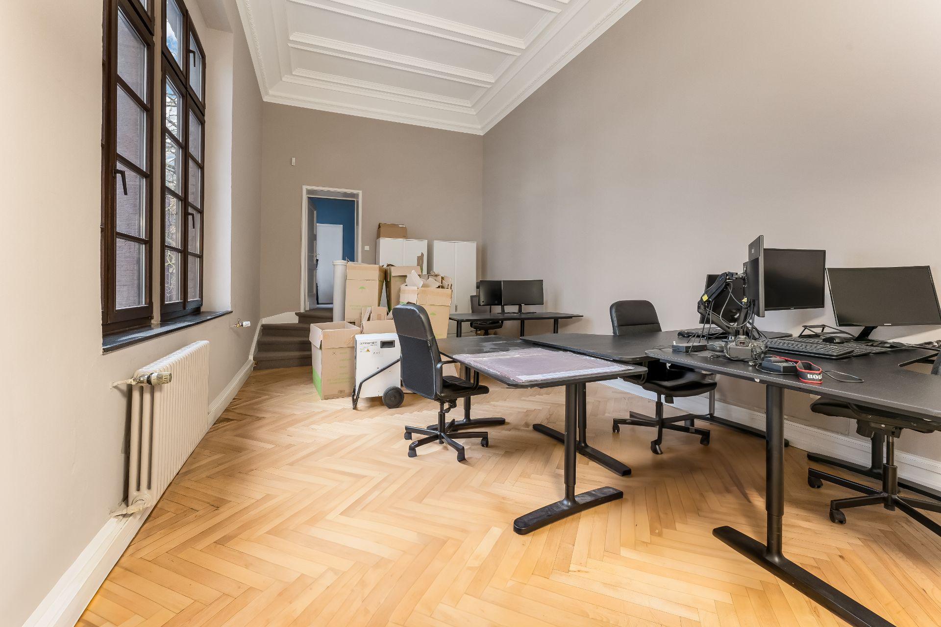 Helles Zimmer mit Fenster, Schreibtischen, Bürostühlen und Umzugskartons auf Parkettboden.