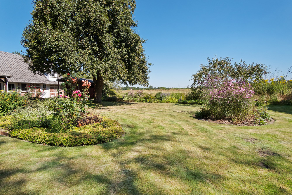 Großer Garten mit Blumenbeeten, Rasen, Bäumen und einem Wohnhaus im Hintergrund.