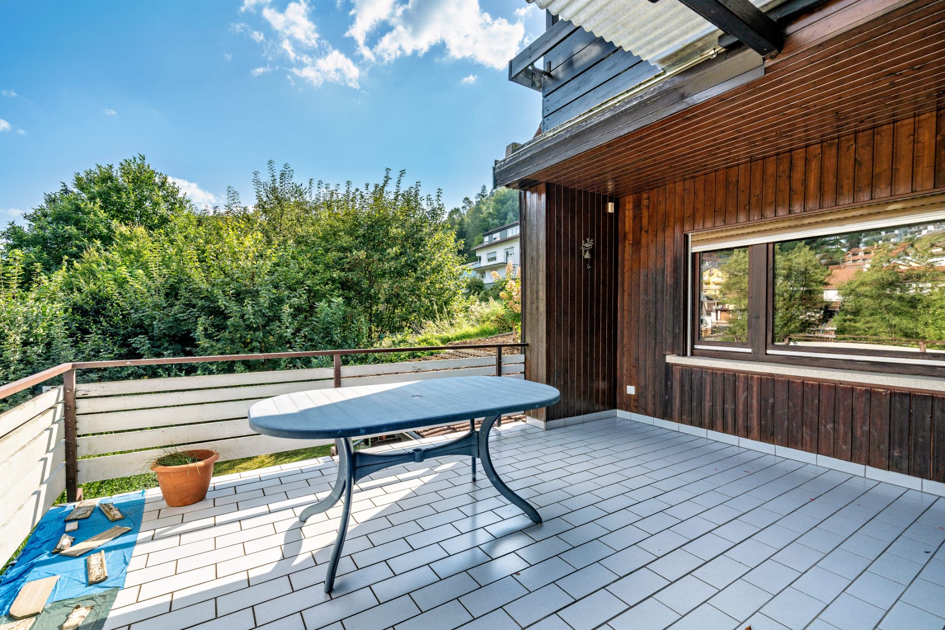 Großer Balkon mit Fliesenboden, blauem Tisch und Blumentopf, umgeben von viel Grün.