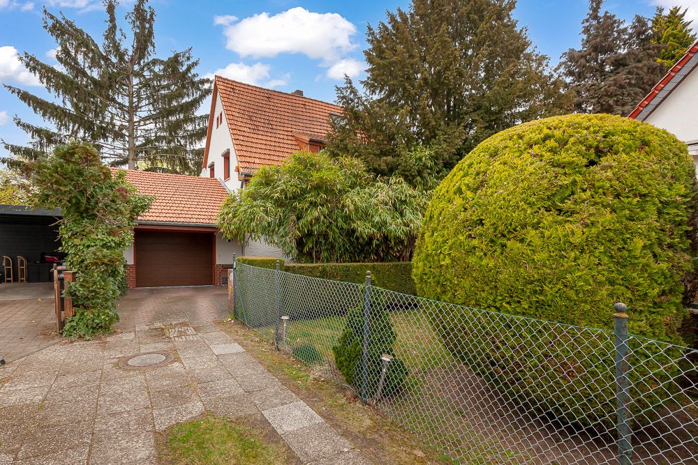 Haus mit rotem Dach, Garage, Garten, Hecke und Metallzaun, umgeben von Bäumen.