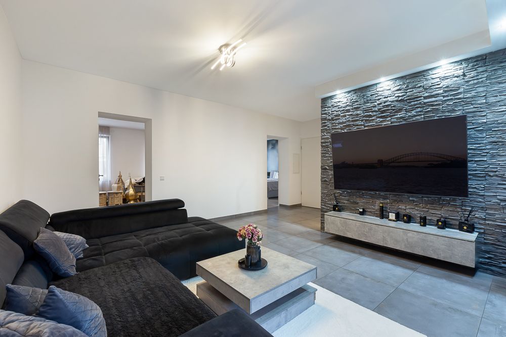 Modernes Wohnzimmer mit schwarzem Sofa, Couchtisch und Wand mit Steinoptik und Fernseher.