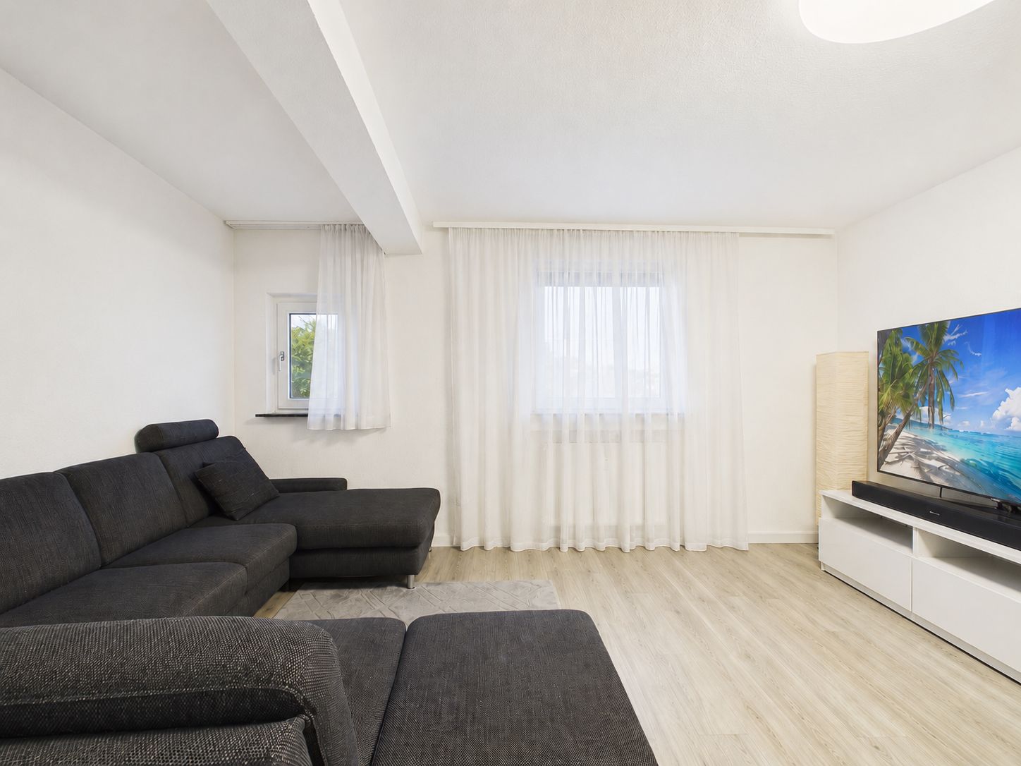 Wohnzimmer mit grauem Ecksofa, Fernseher auf TV-Schrank und hellem Holzboden.