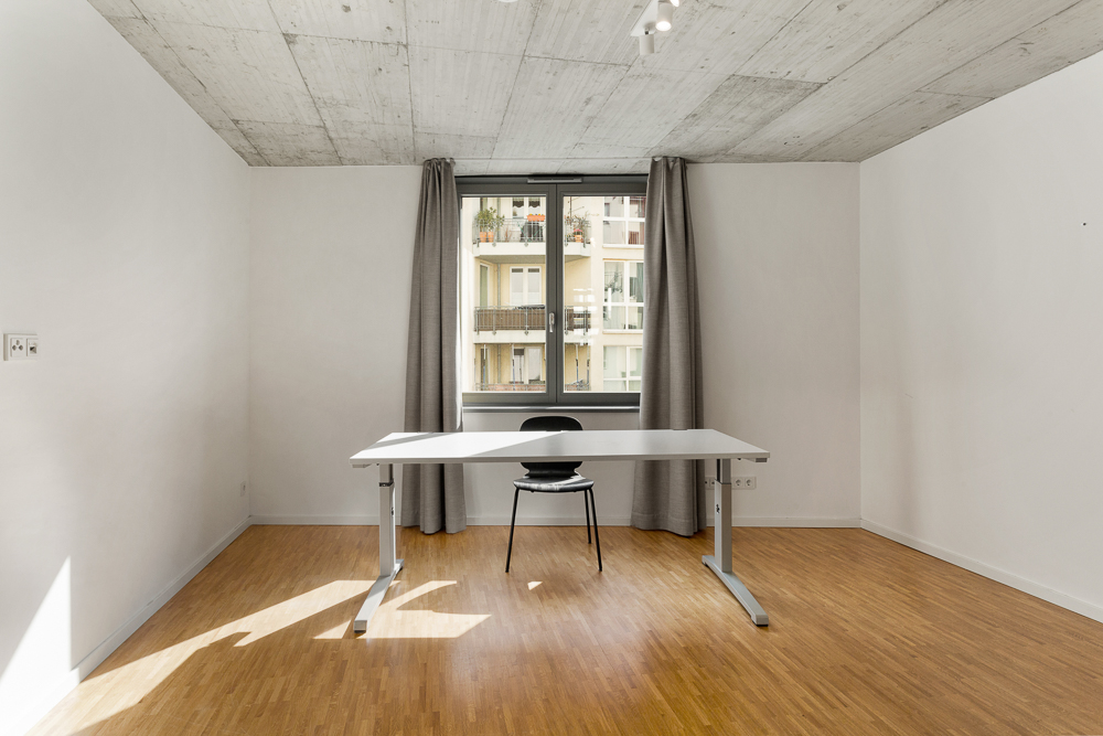 Leerer Raum mit Schreibtisch, Stuhl, Holzfußboden und Fenster mit grauen Vorhängen.