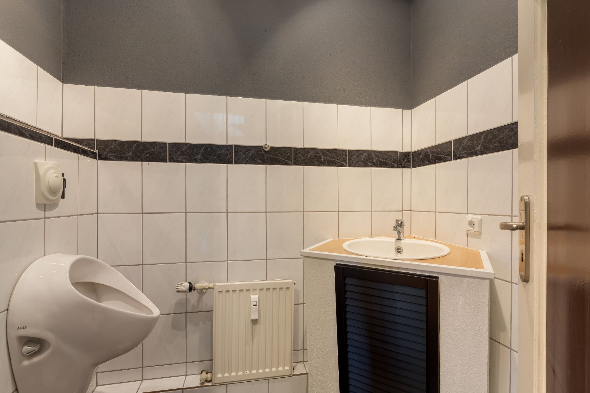 Kleines Badezimmer mit Urinal, Waschbecken und Heizkörper, weiße Fliesen an den Wänden.