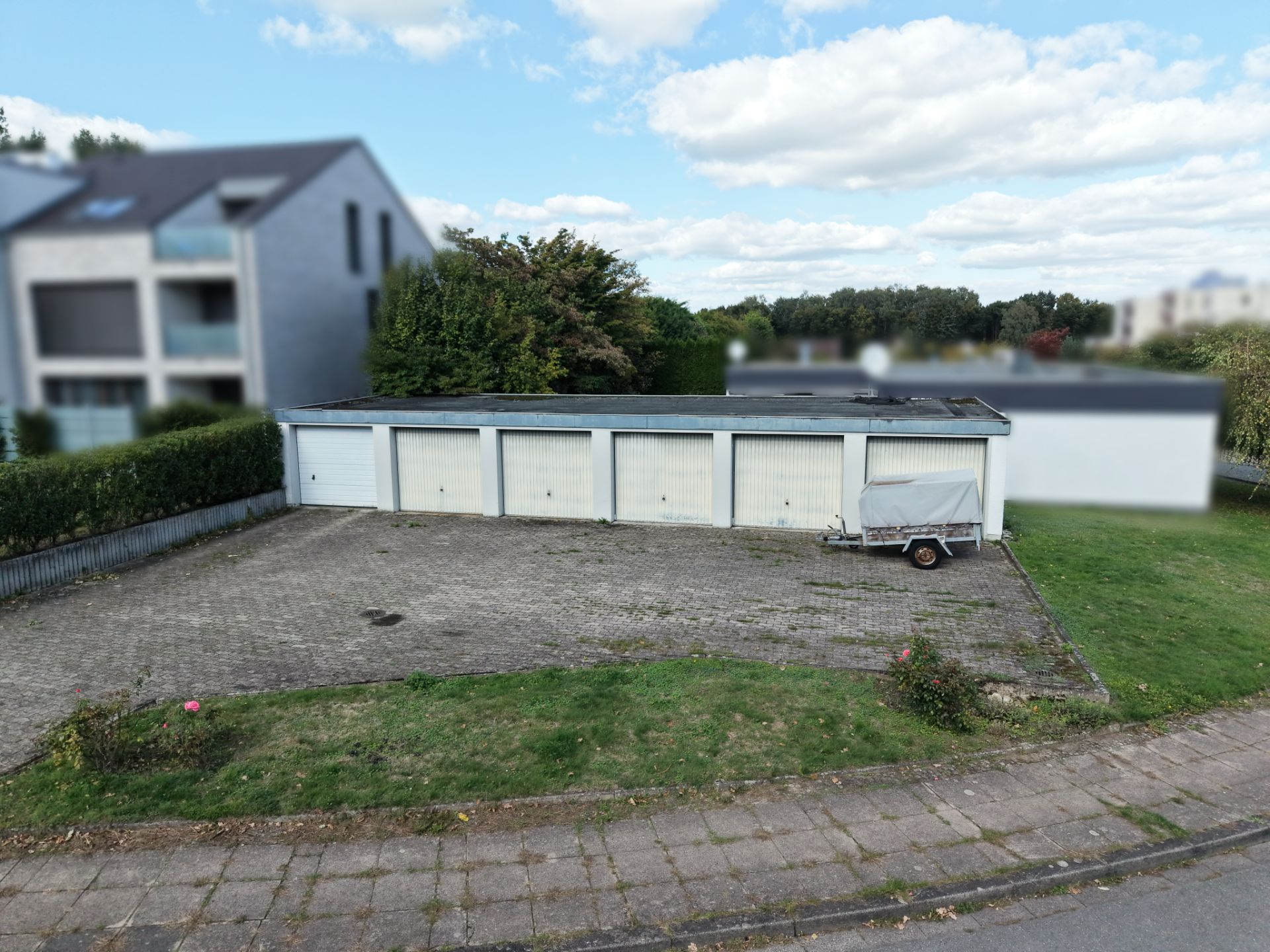 Sechs Garagen mit weißem Tor und ein Anhänger auf einem gepflasterten Hof, daneben Rasenfläche.