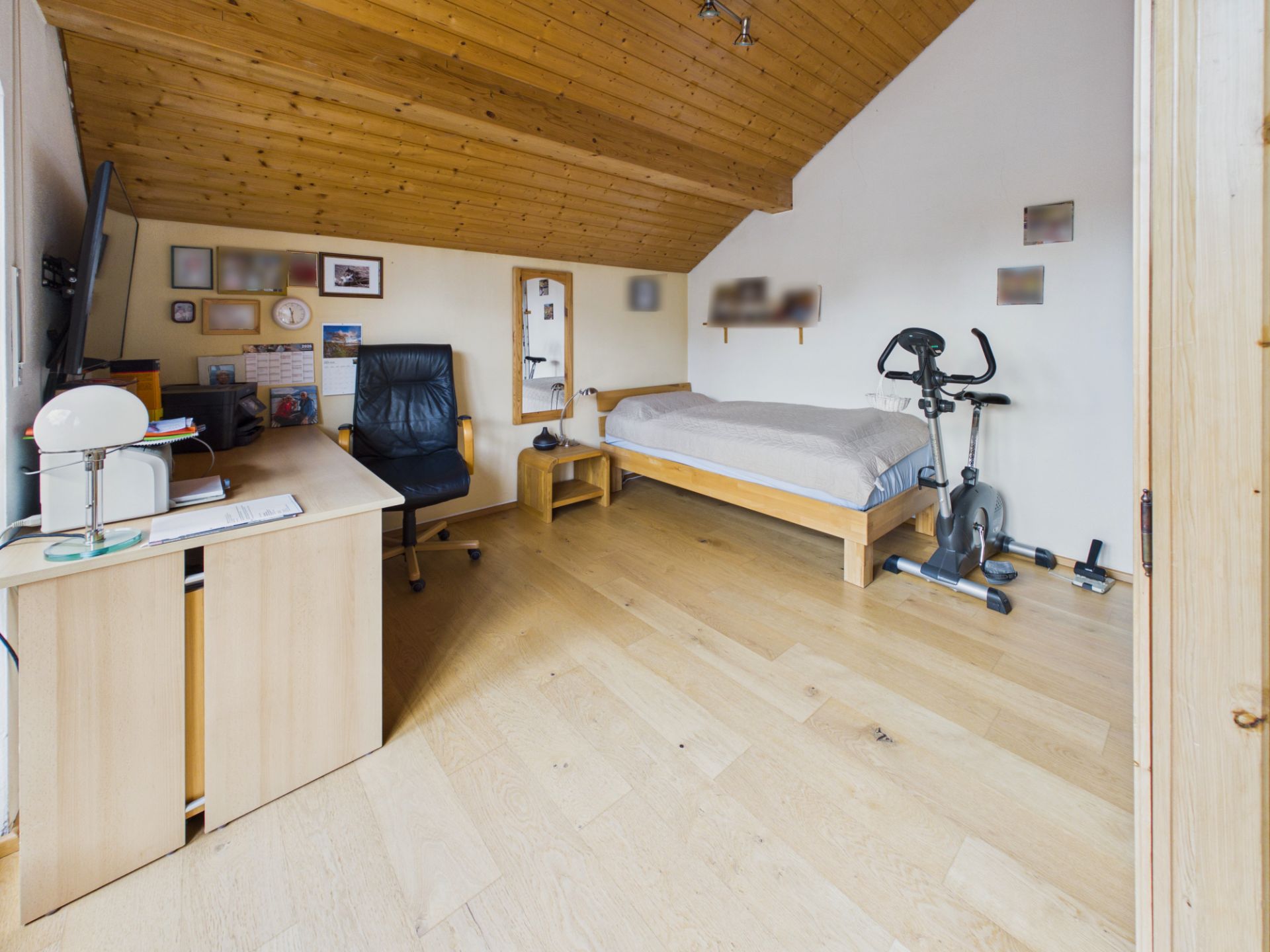 Zimmer mit Schreibtisch, Bürostuhl, Bett und Heimtrainer unter einer Holzdecke.