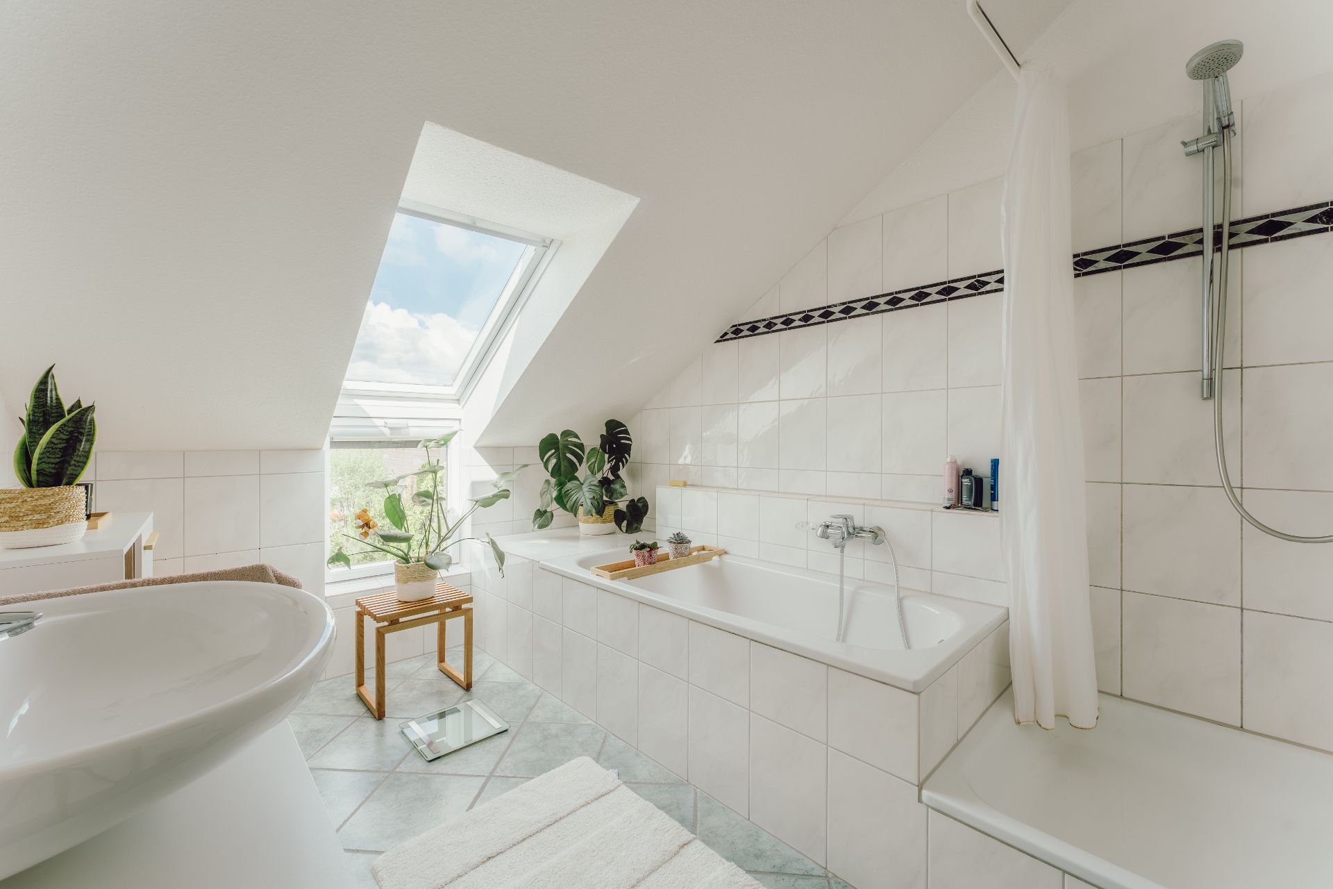 Helles Badezimmer mit Badewanne, Dachfenster, Pflanzen und weiß gefliesten Wänden.