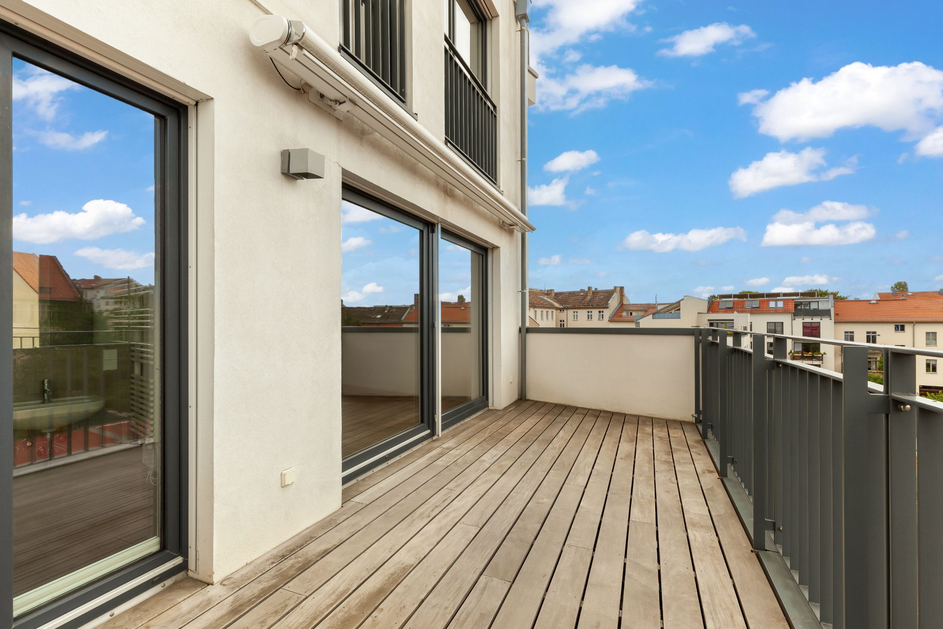 Großer Balkon mit Holzboden, Glasgeländer und Blick auf Nachbargebäude unter blauem Himmel.