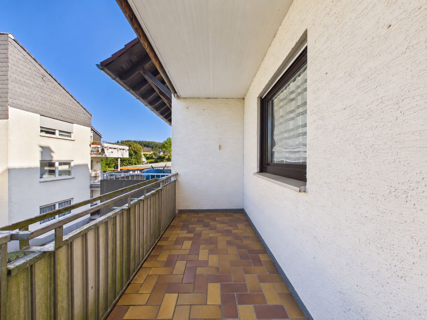 Balkon mit braunem Fliesenboden, Metallgeländer und Blick auf Nachbarhäuser.