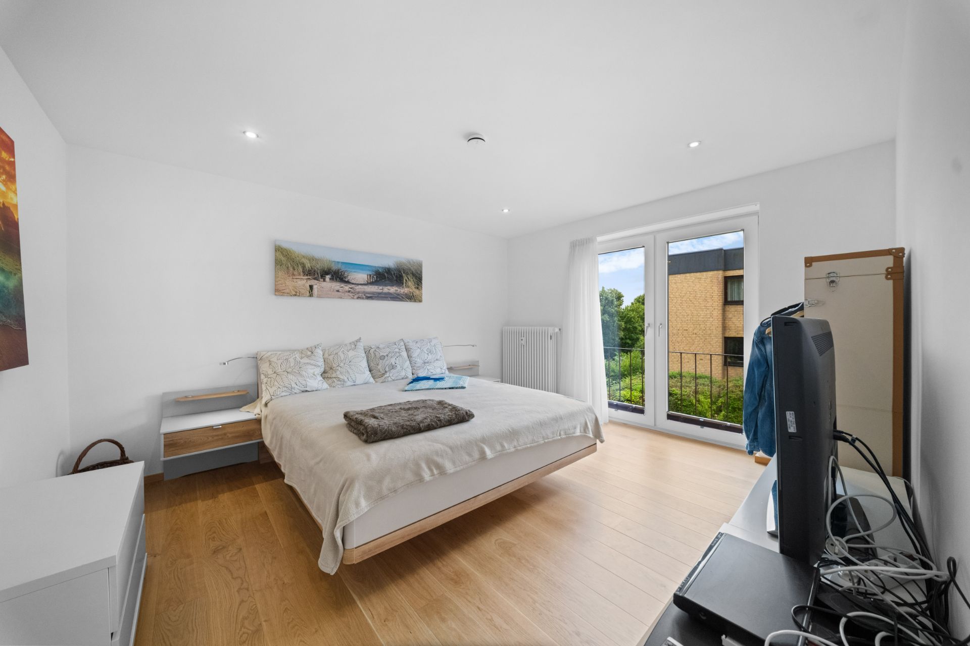 Helles Schlafzimmer mit Doppelbett, Fenster, Fernseher und Holzboden.