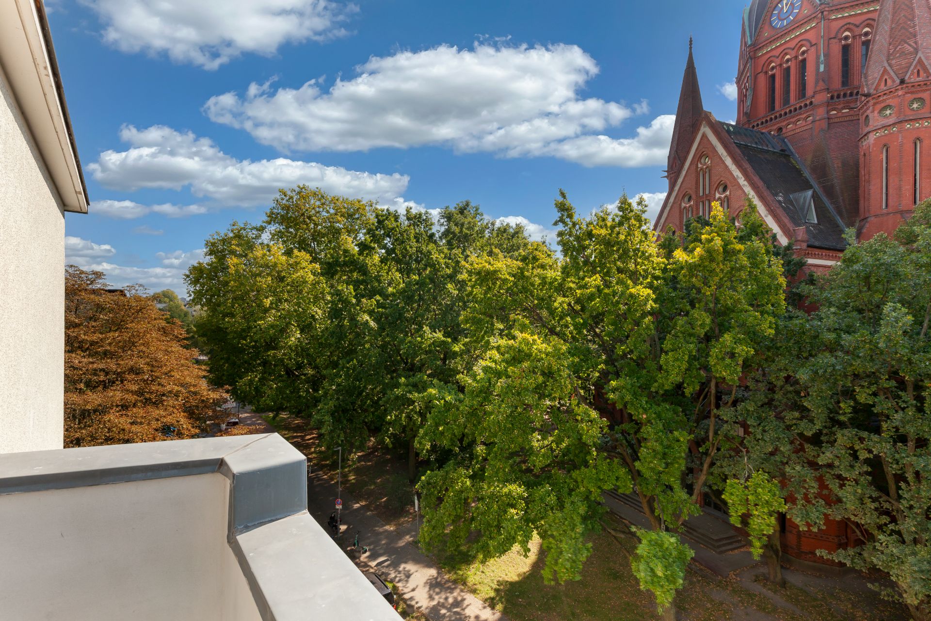 Blick von einem Balkon auf Bäume und ein großes rotes Kirchengebäude bei sonnigem Wetter.