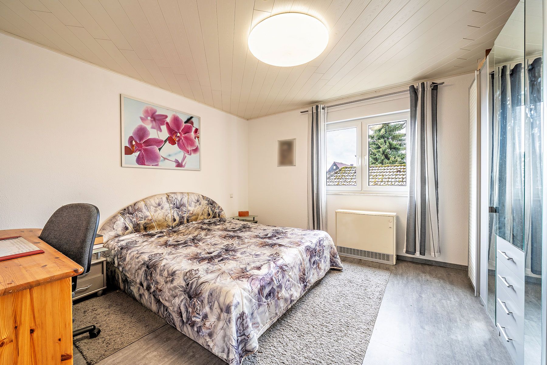 Helles Schlafzimmer mit Doppelbett, Schreibtisch, Fenster und Bild mit rosa Orchideen an der Wand.