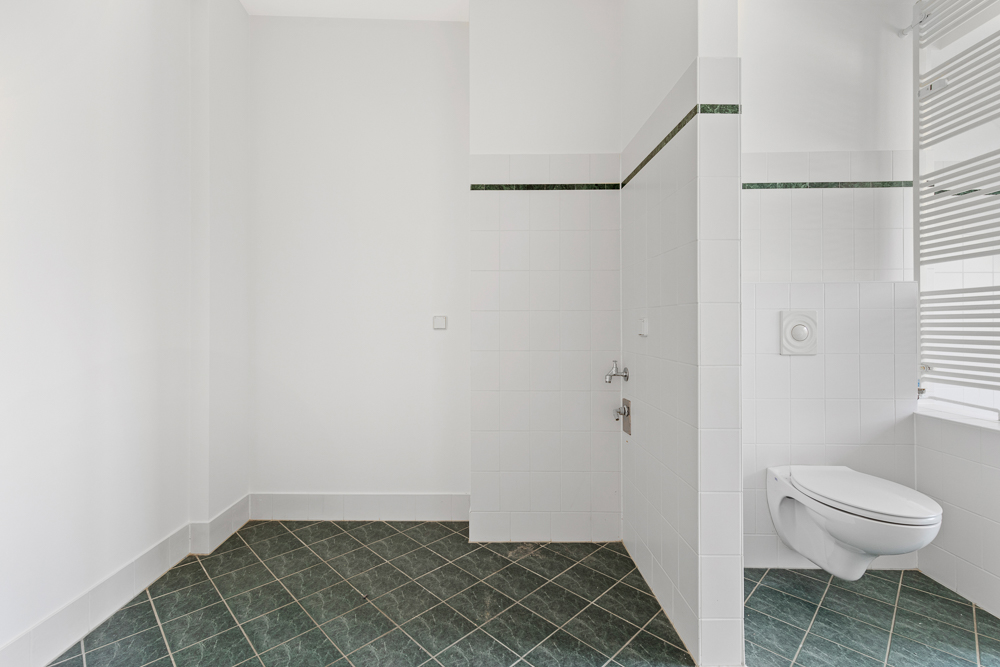 Helles Badezimmer mit grünen Fliesen und Wand-WC, rechteckigem Fenster mit Jalousien.