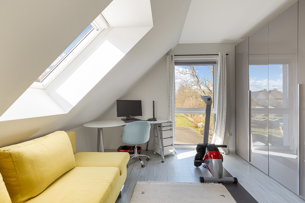 Helles Zimmer mit gelbem Sofa, Schreibtisch, Computer, Heimtrainer und großen Fenstern.