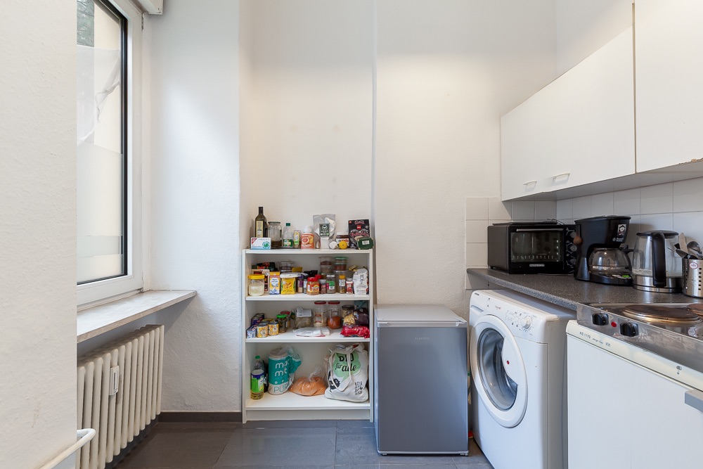 Kleine Küche mit Fenster, Regal voller Lebensmittel, Kühlschrank, Waschmaschine und Küchengeräten auf der Arbeitsplatte.