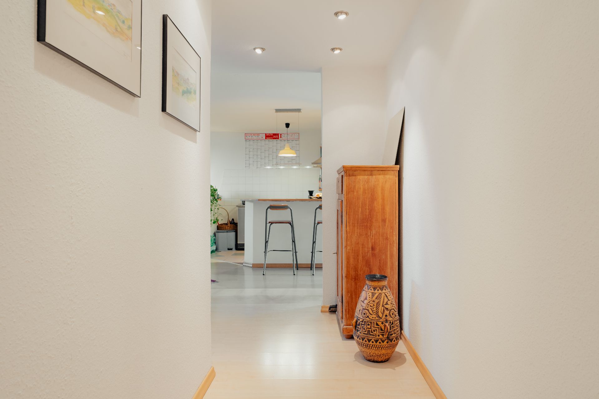 Heller Flur mit Holzschrank, Vase und Bildern an den Wänden, Blick in eine moderne Küche.