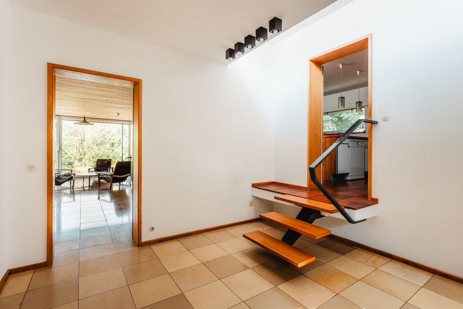 Flur mit Treppe, Blick in ein Wohnzimmer mit Sesseln und Fensterfront, rechts Zugang zur Küche.