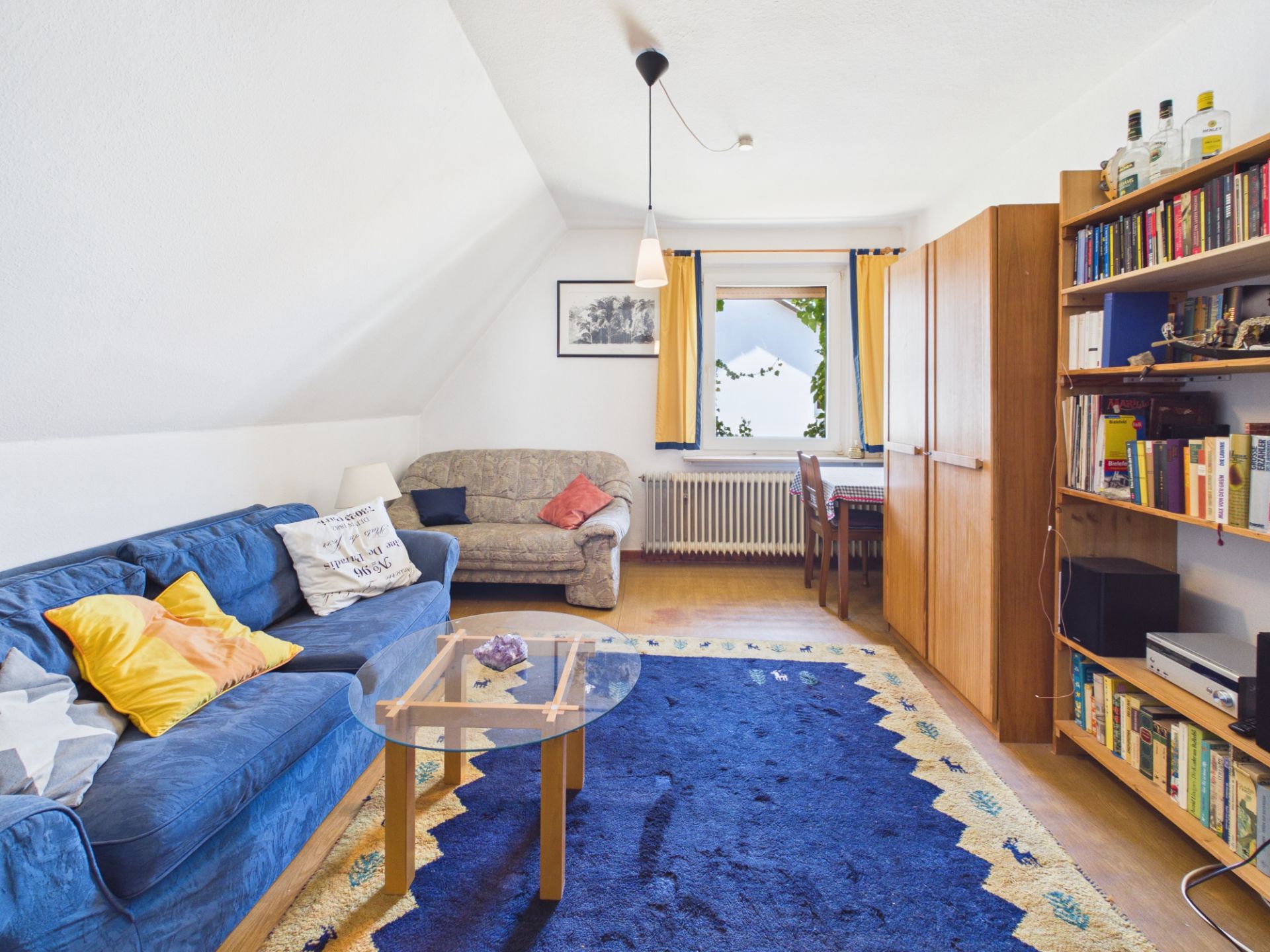 Wohnzimmer mit blauem Sofa, Teppich, Couchtisch, Bücherregal, Sessel und Fenster mit gelben Vorhängen.