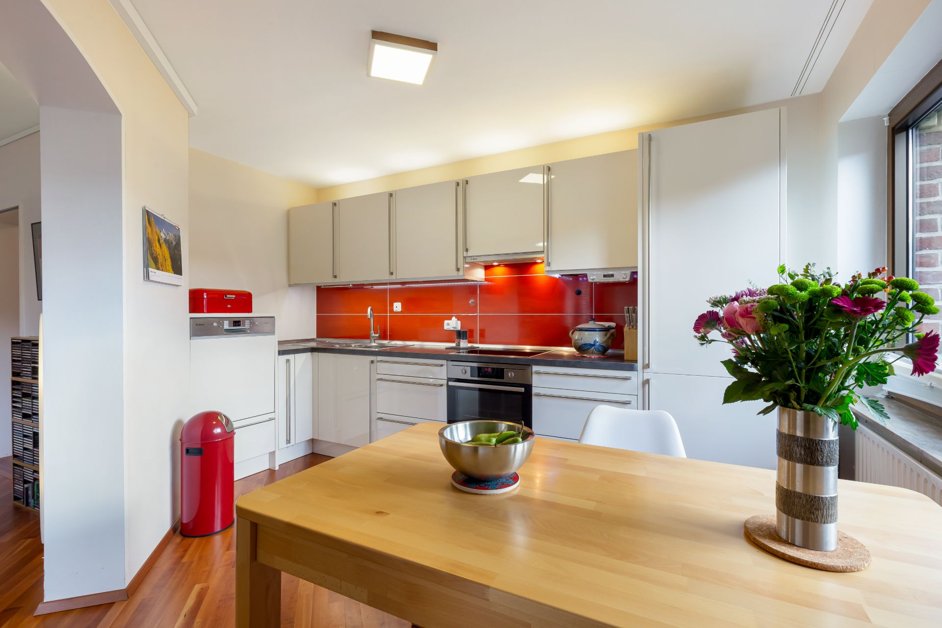 Helle Küche mit weißem Mobiliar, roter Rückwand, Holztisch, Schale und Blumenstrauß.