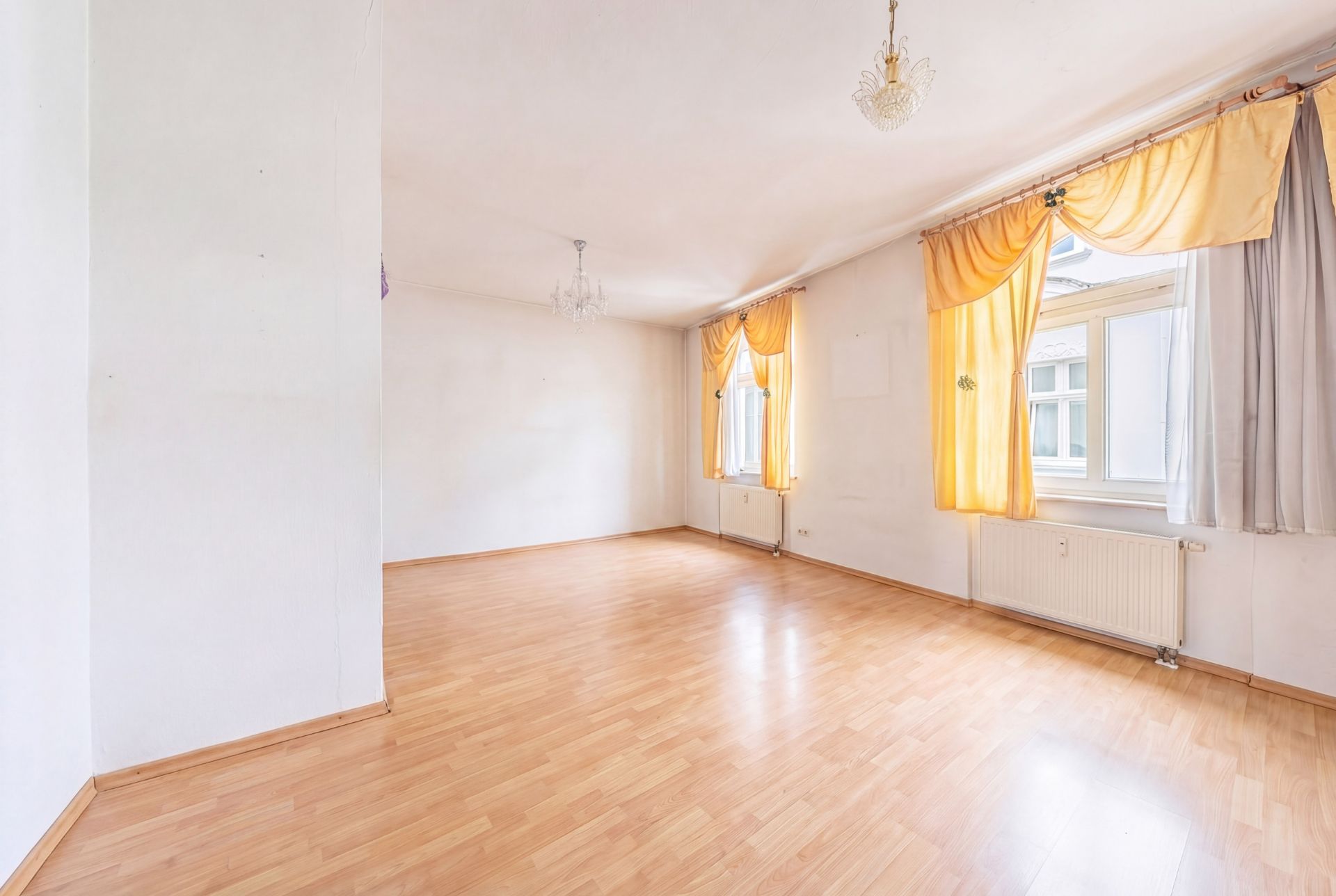 Heller, leerer Raum mit Holzfußboden, zwei Fenstern und gelben Vorhängen.