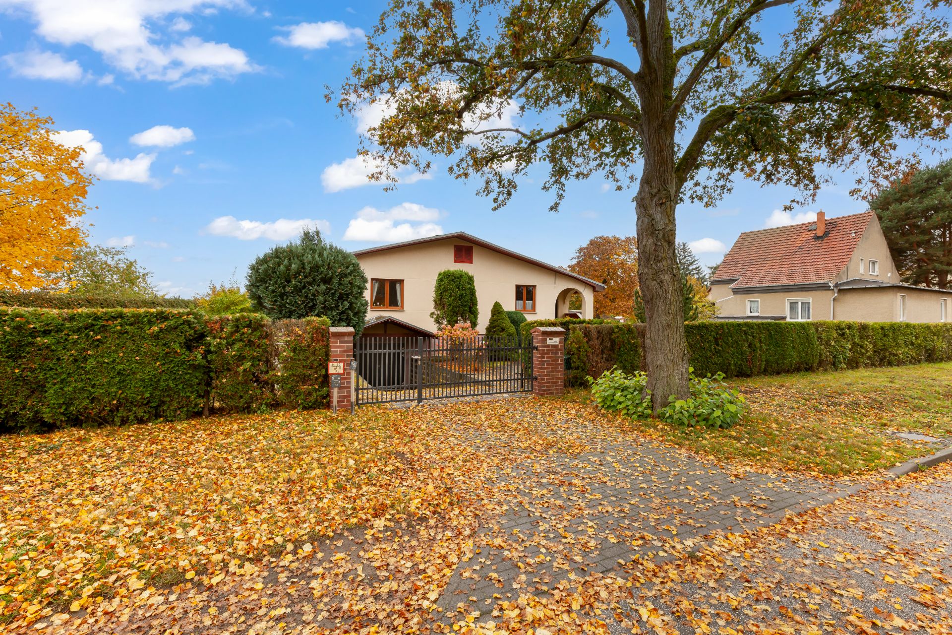 Einfamilienhaus mit Garten, Einfahrtstor und Baum, Herbstlaub auf dem Boden.