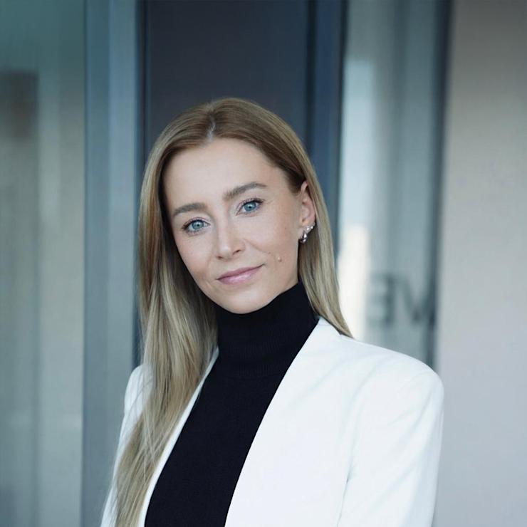 Frau mit langen blonden Haaren im weißen Blazer steht vor Glaswand.
