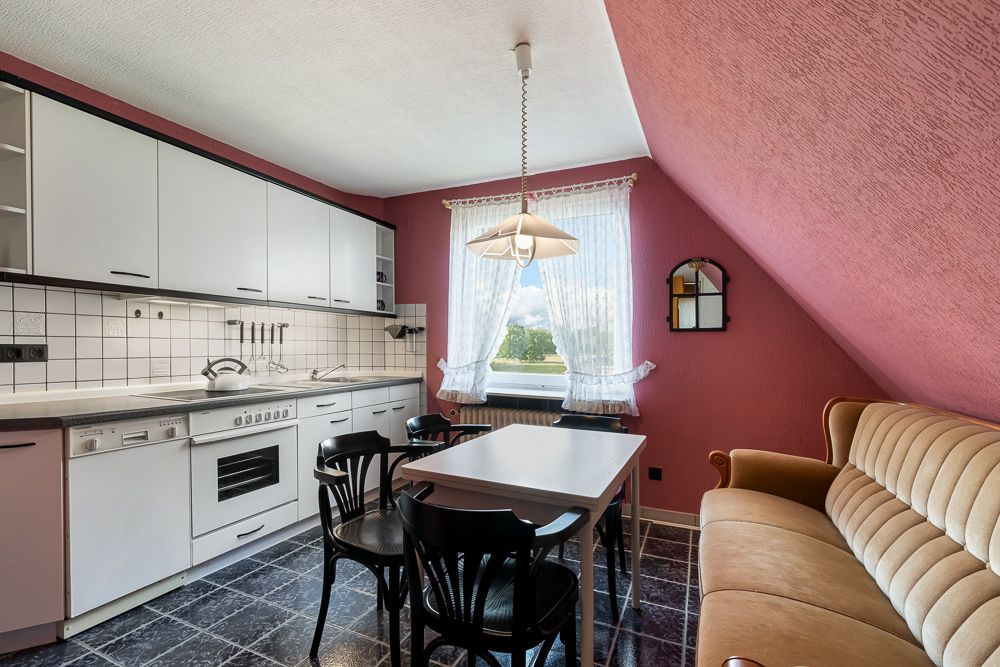 Küche mit weißen Schränken, Esstisch, vier Stühlen, beige Sofa und Fenster mit Vorhängen.