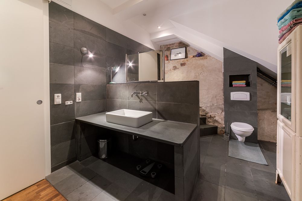 Modernes Badezimmer mit dunklen Fliesen, Waschbecken, Spiegel und Toilette unter einer Dachschräge.