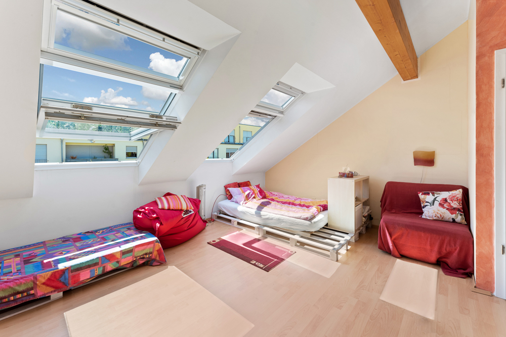 Dachzimmer mit zwei Betten, Sessel, buntem Teppich und großen Dachfenstern.