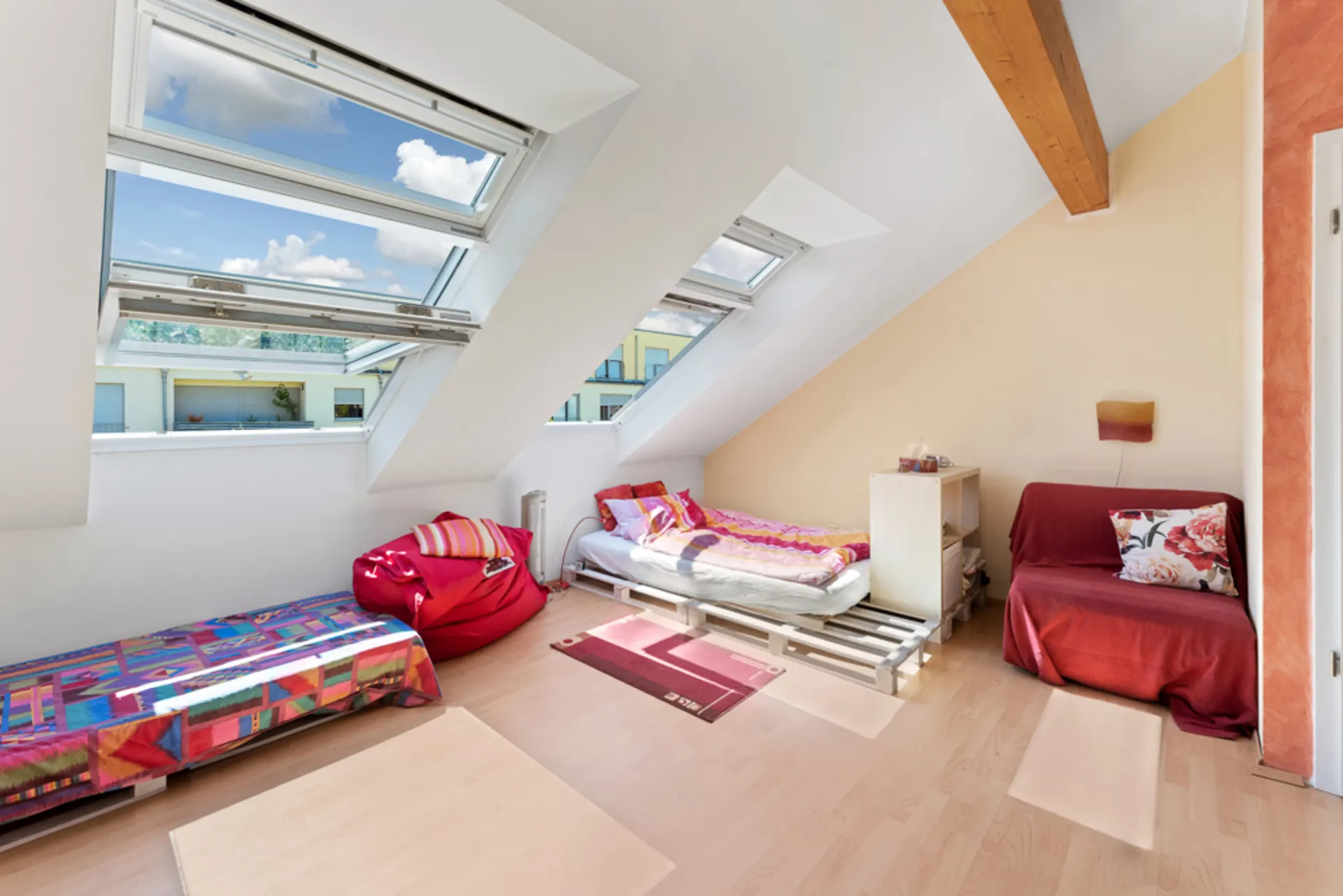 Dachzimmer mit zwei Betten, Sessel, buntem Teppich und großen Dachfenstern.