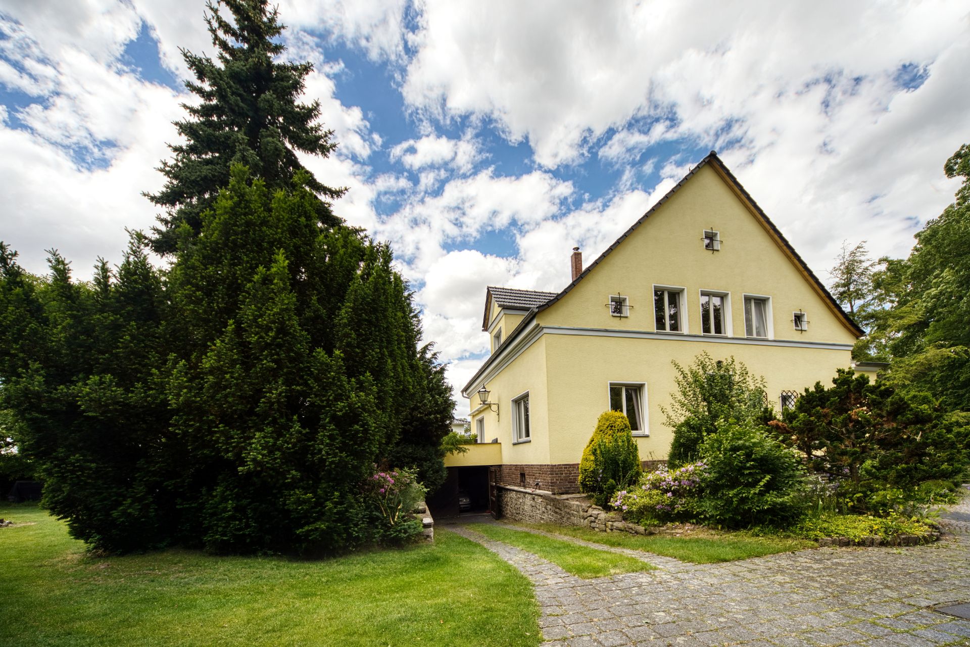 Gelbes Einfamilienhaus mit Garten, Bäumen und gepflastertem Weg bei bewölktem Himmel.