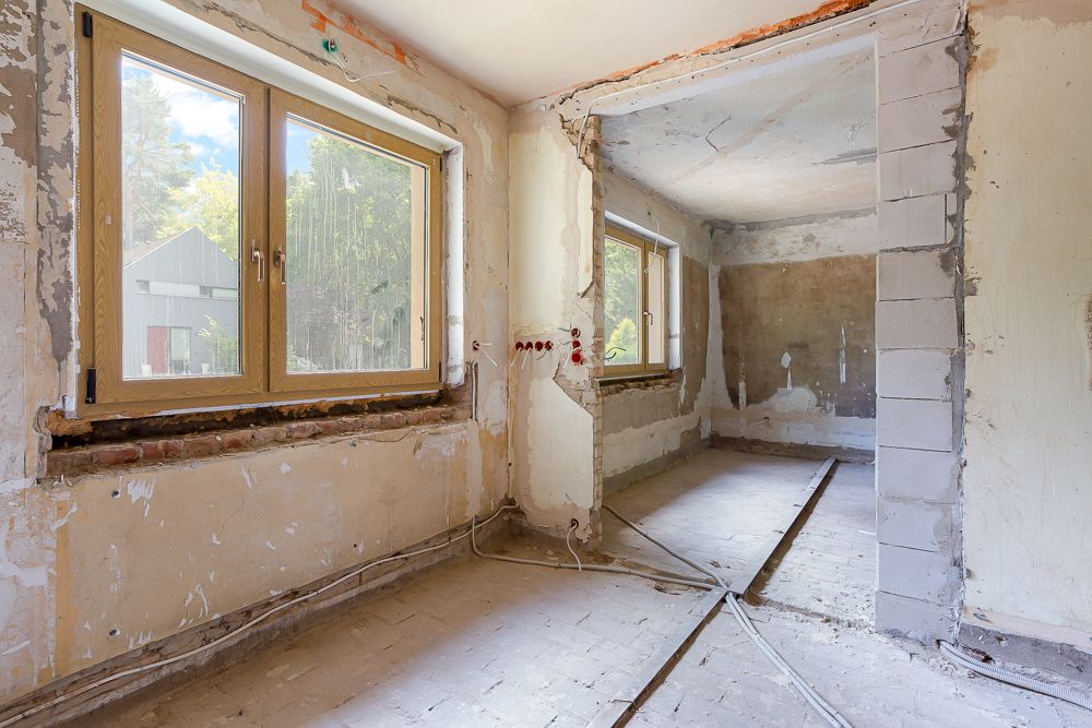 Zimmer eines Hauses im Rohbau mit großen Fenstern und sichtbaren Leitungen.