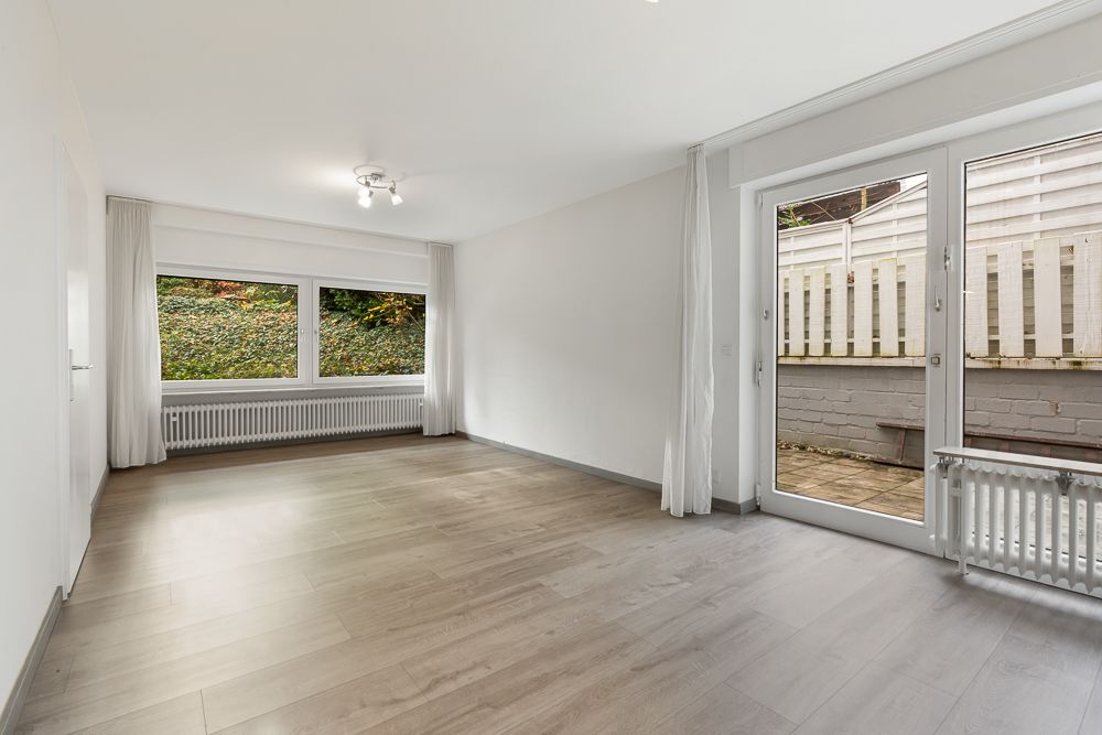Helles, leeres Wohnzimmer mit Holzfußboden, großen Fenstern und Zugang zur Terrasse.