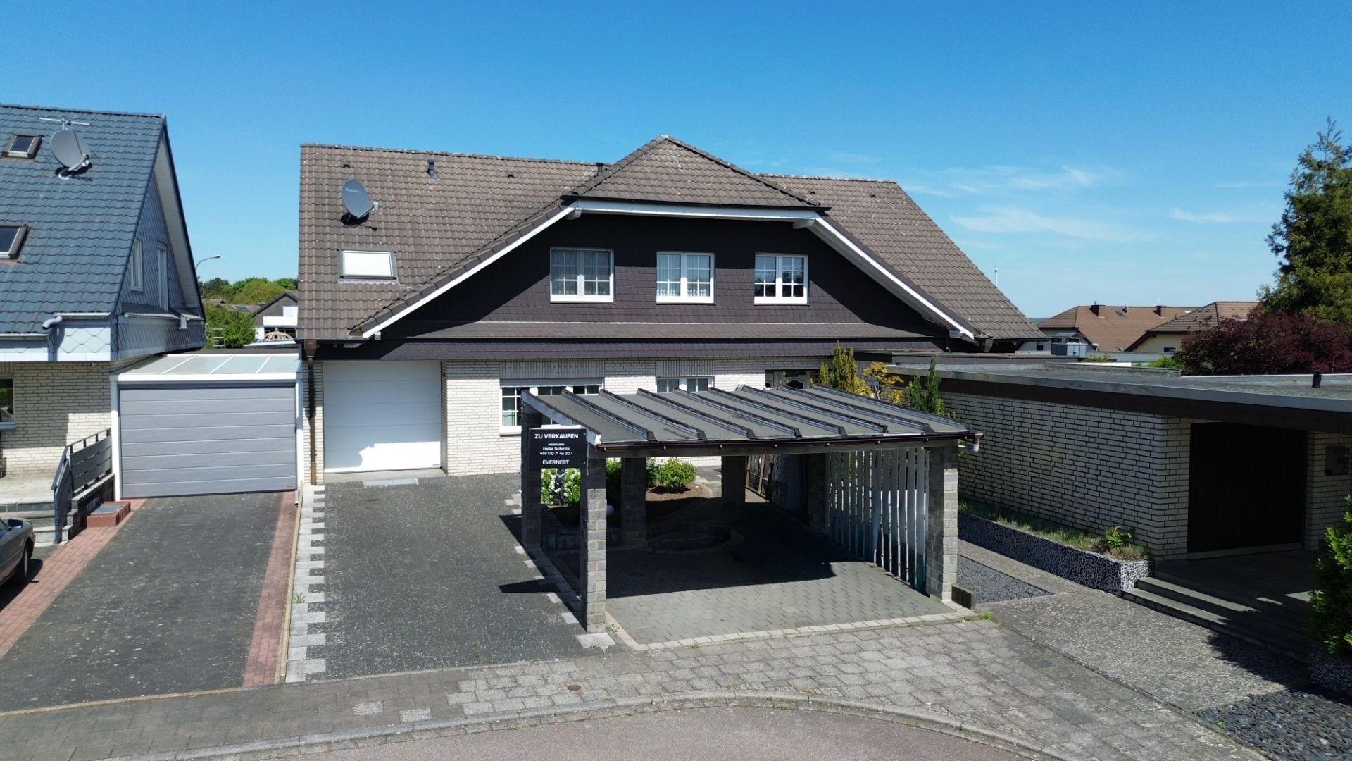 Zweistöckiges Wohnhaus mit Satteldach, Carport und gepflasterter Einfahrt bei klarem Himmel.