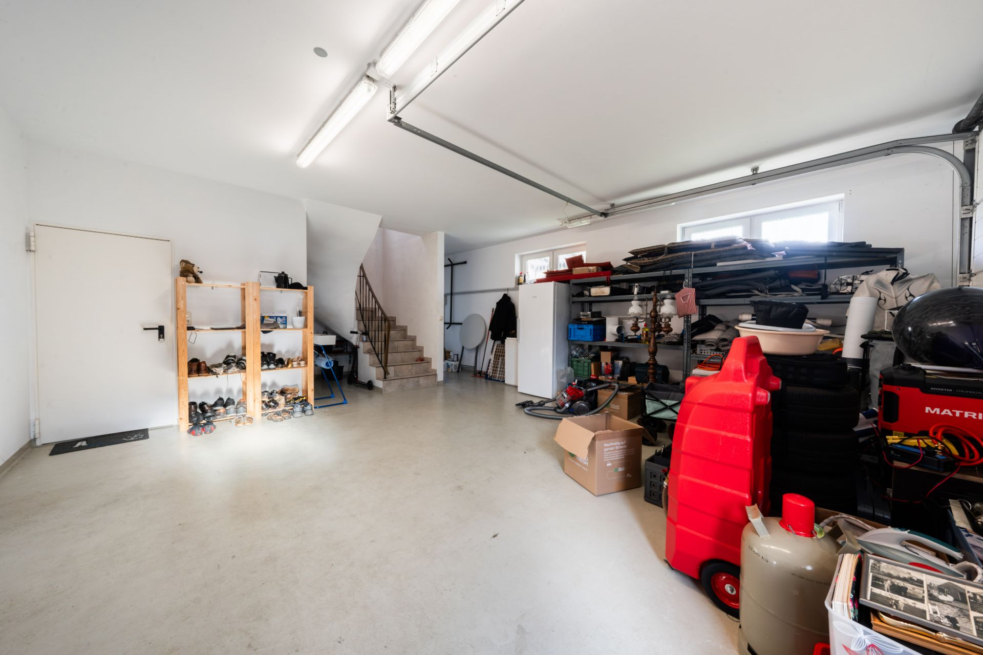 Aufgeräumte Garage mit Regalen, Schuhen, Kartons und einer Treppe im Hintergrund.