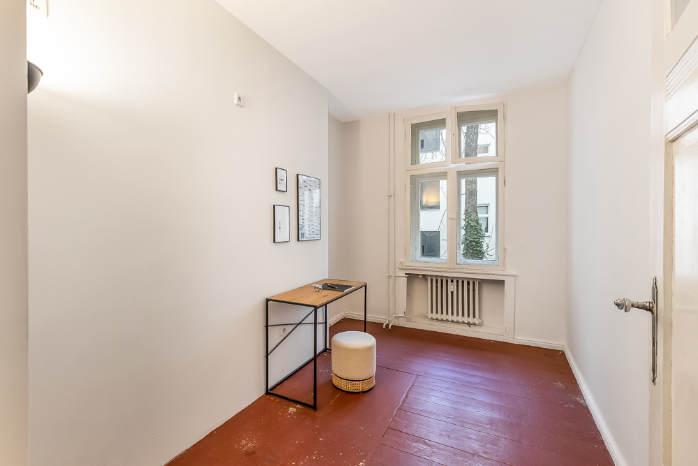 Kleines Zimmer mit Schreibtisch, Hocker, Wandbildern und großem Fenster mit Heizkörper.