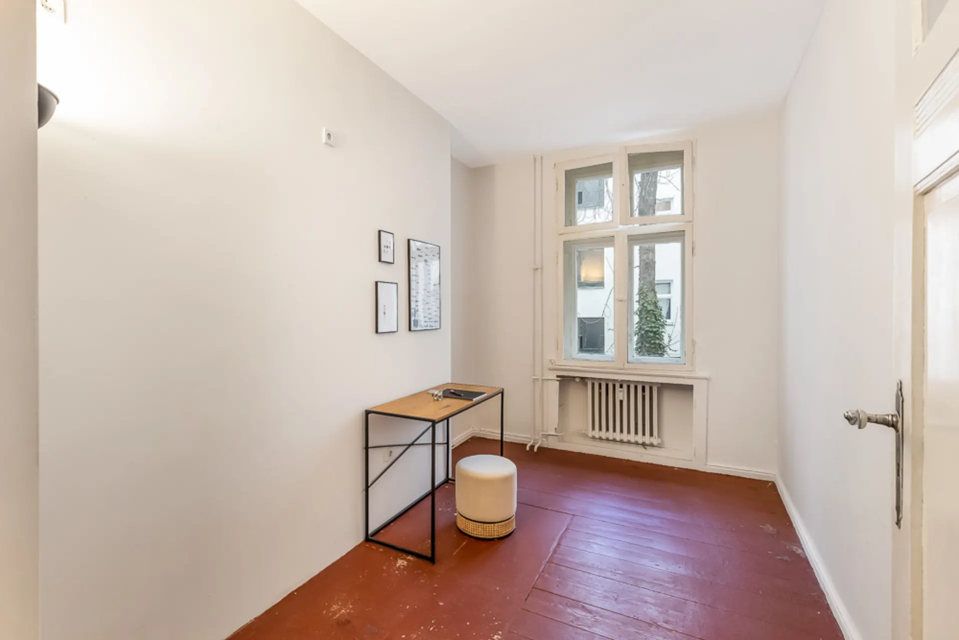 Kleines Zimmer mit Schreibtisch, Hocker, Wandbildern und großem Fenster mit Heizkörper.