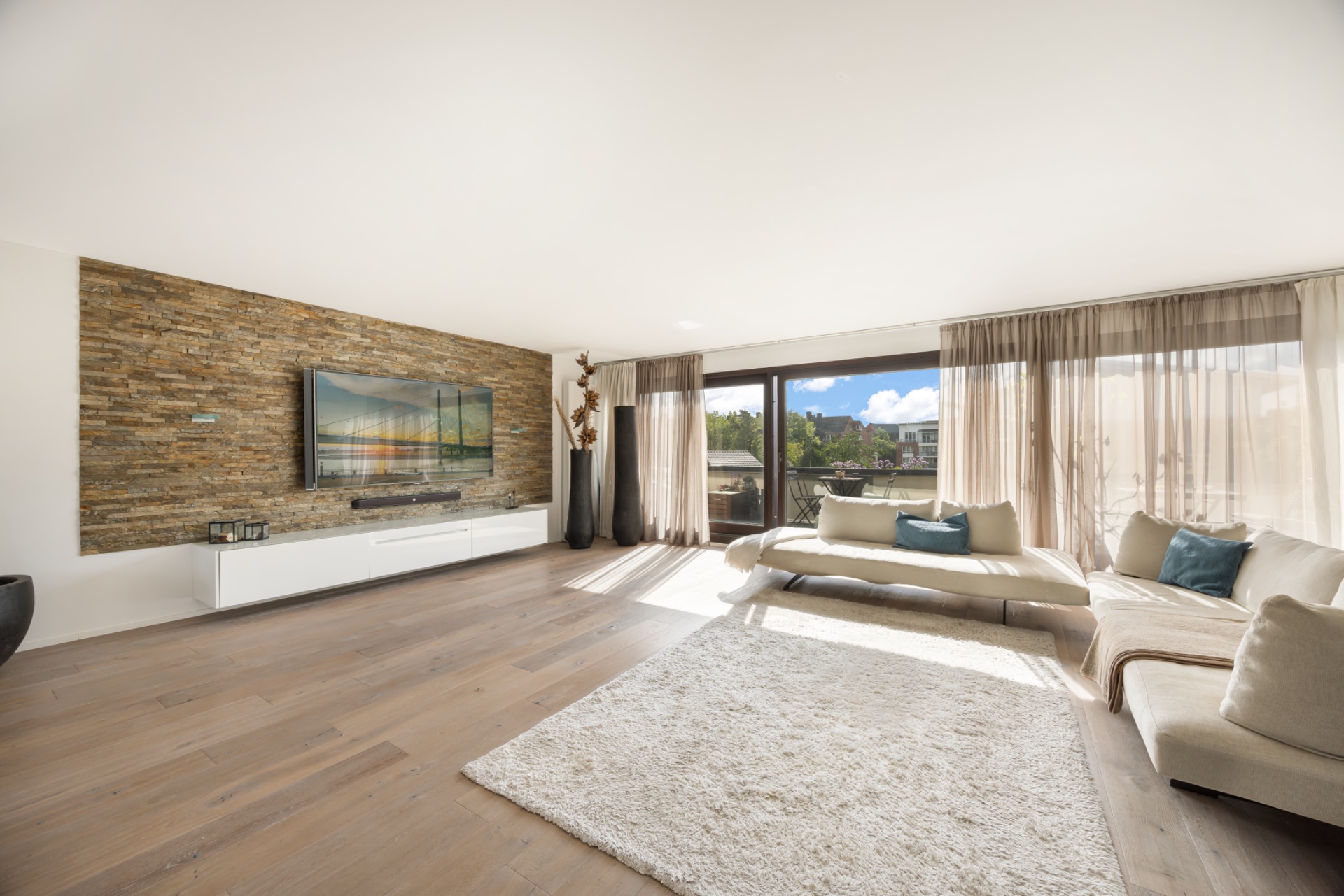 Helles Wohnzimmer mit Holzfußboden, großer Fensterfront, Sofas und Wand mit Fernseher.