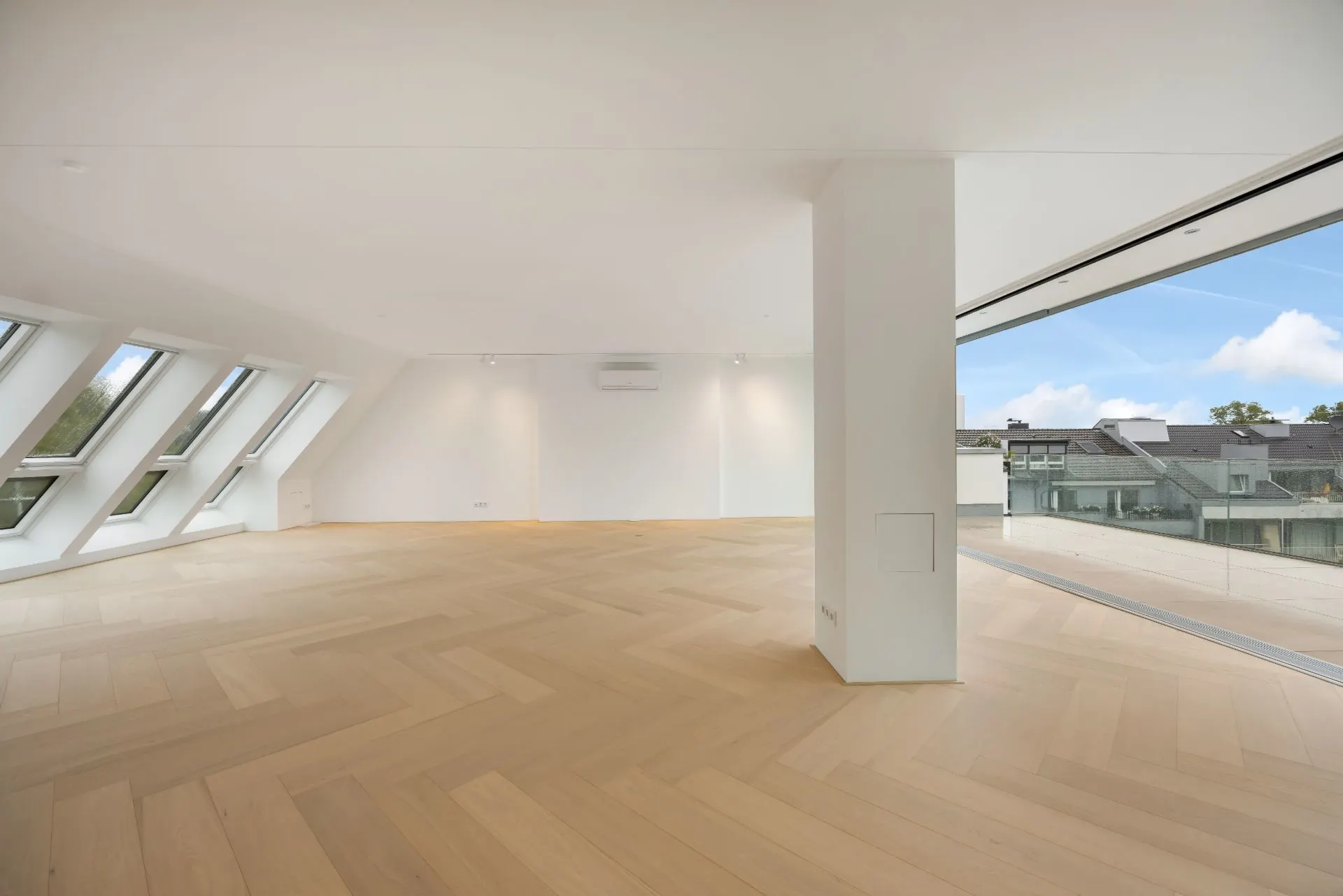 Helles, leeres Zimmer mit Holzfußboden, schrägen Fenstern und großer Fensterfront.