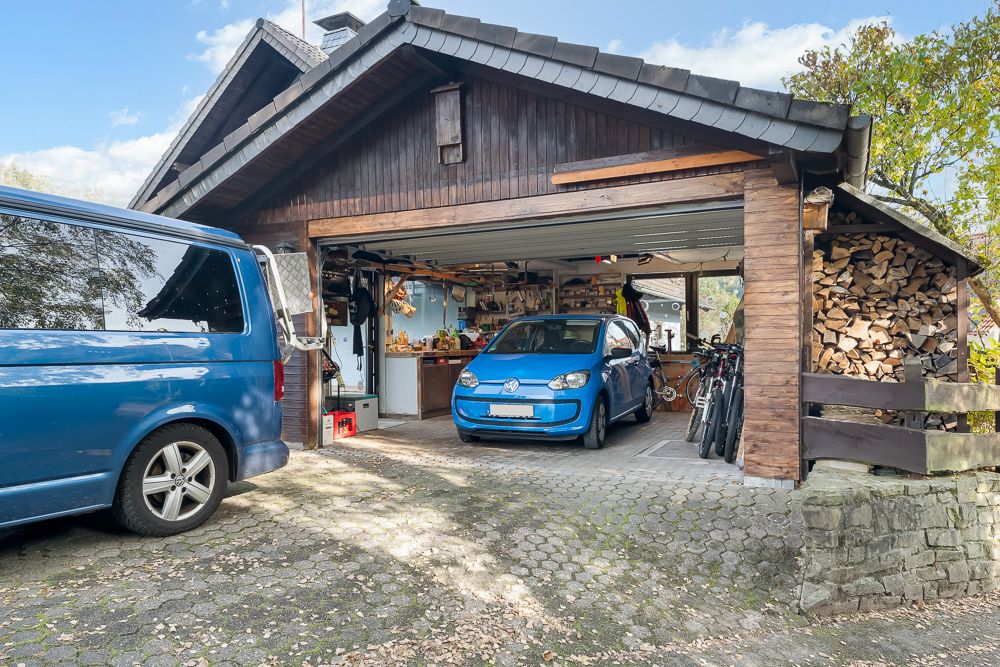 Holzgarage mit offenem Tor, zwei Autos, Fahrräder und gestapeltes Brennholz sichtbar.