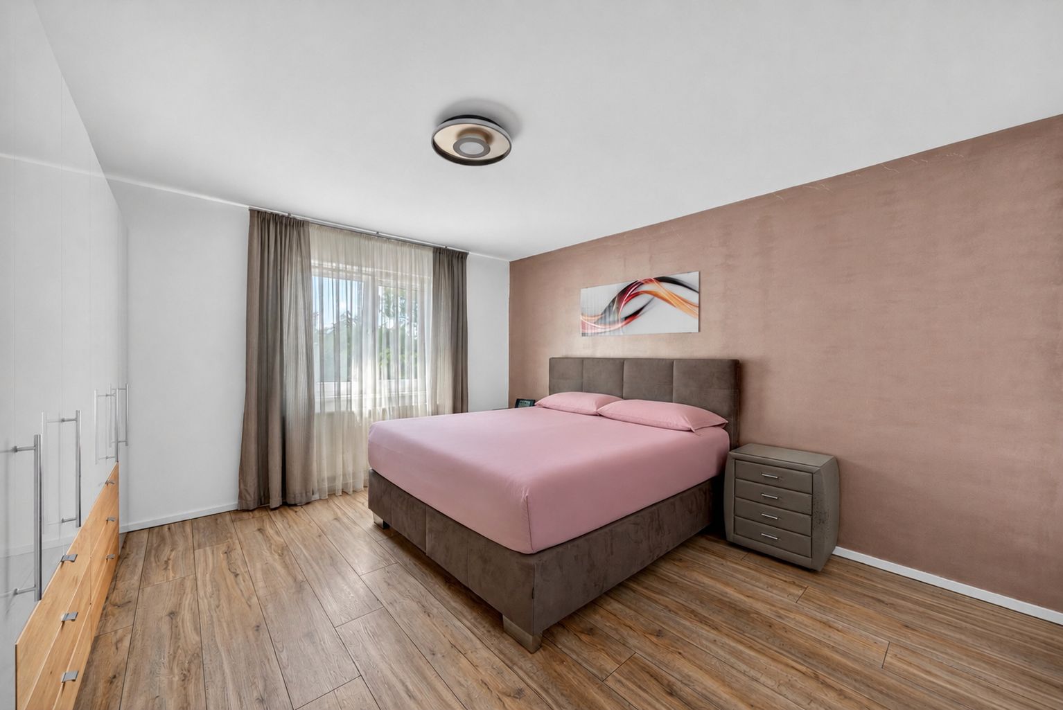 Schlafzimmer mit Doppelbett, rosa Bettwäsche, Nachttisch und großem Fenster mit Vorhängen.