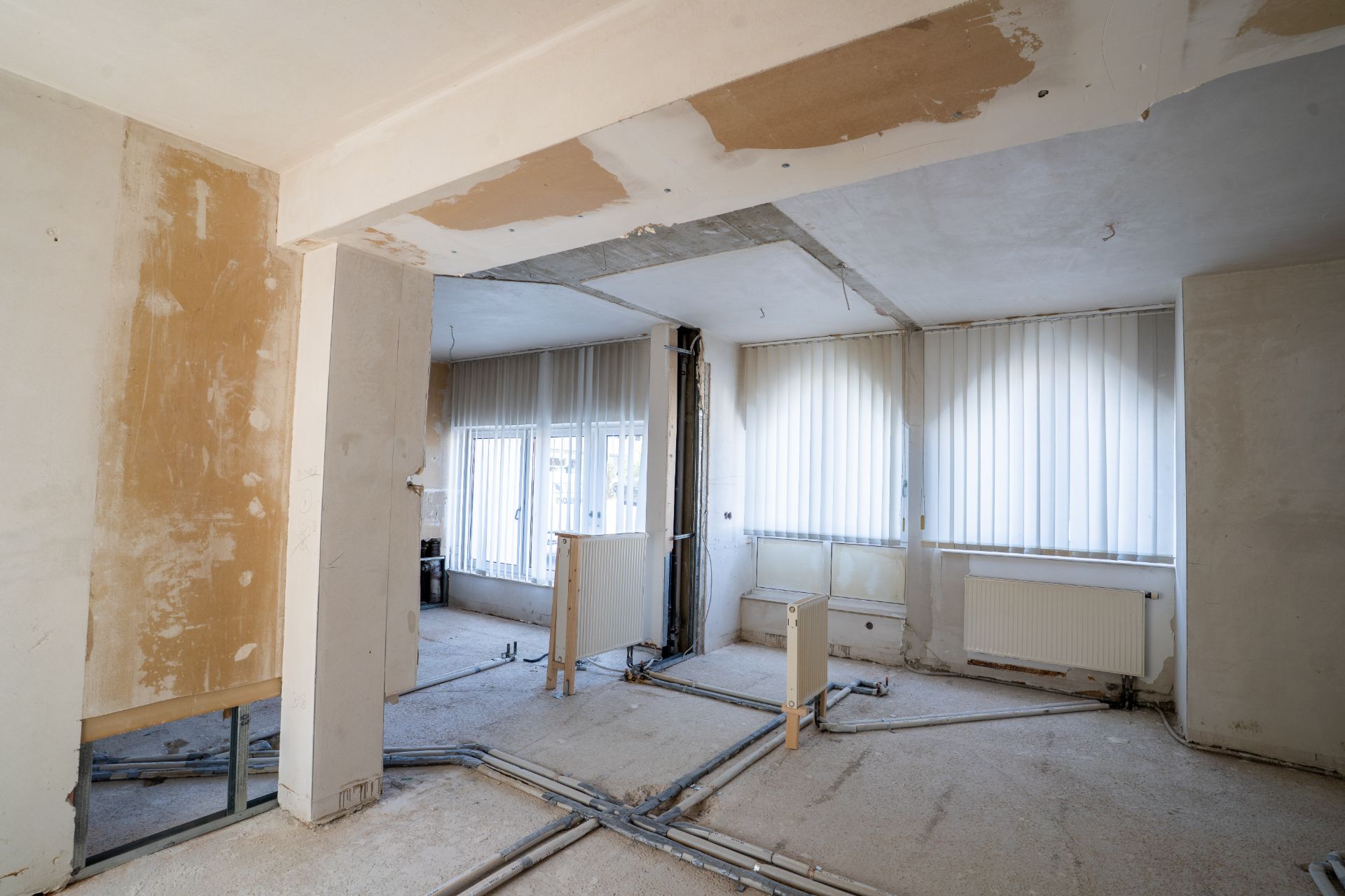 Leerer Raum im Umbau mit freiliegenden Leitungen und großen Fenstern mit Jalousien.